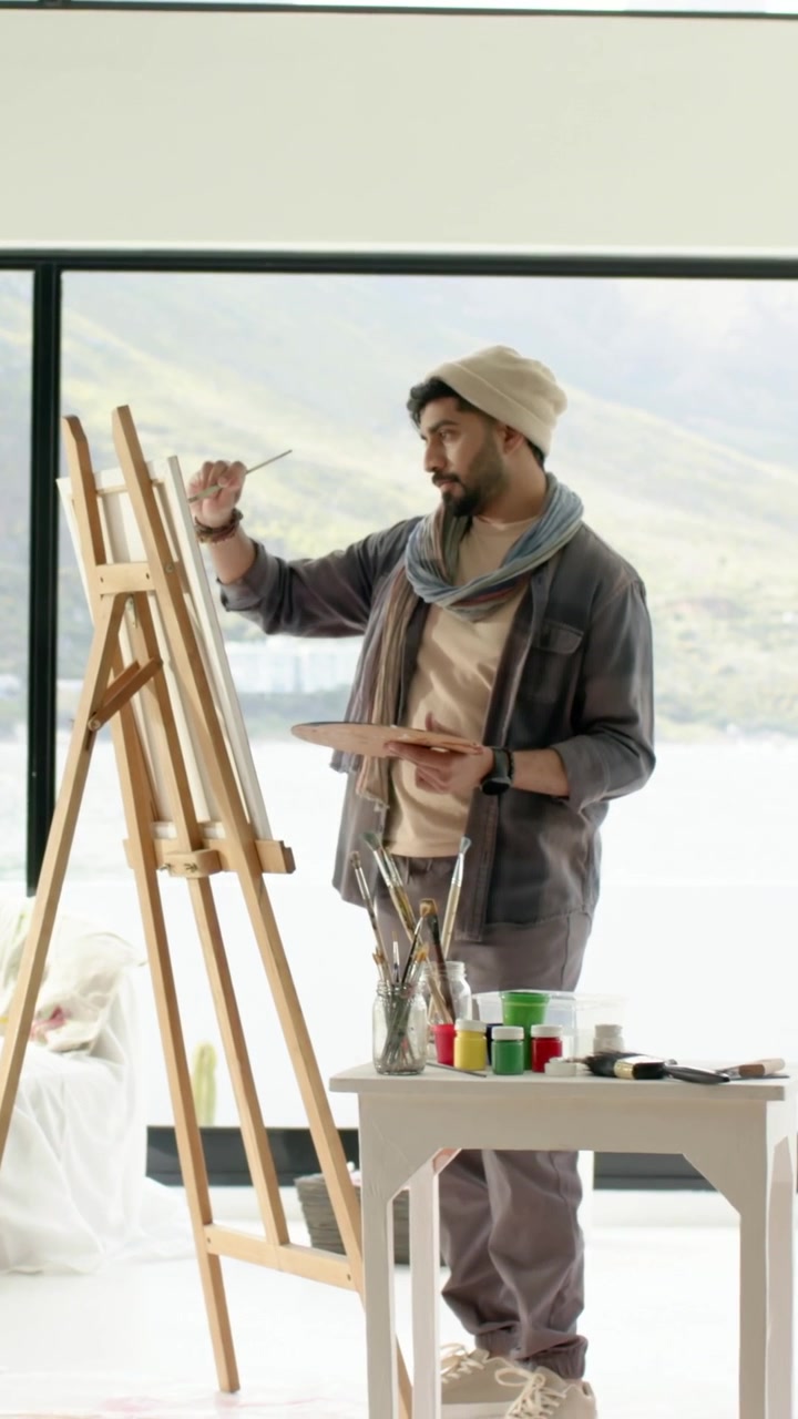 Dikey video: Beresi ve atkısıyla, paletiyle pencerenin yanında manzara resmi yapan, fırçasını kaldıran ressam.