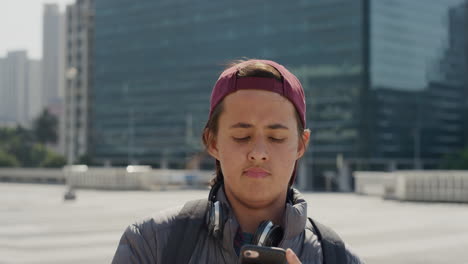 portrait relaxed young teenage boy using smartphone texting browsing online messages on mobile phone looking pensive in sunny urban city slow motion