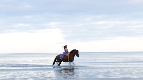 Una-Hermosa-Chica-Con-El-Pelo-Largo-Y-Un-Vestido-Azul-Montando-A-Caballo-Por-El-Agua-Durante-La-Noche,-Donabate,-Irlanda