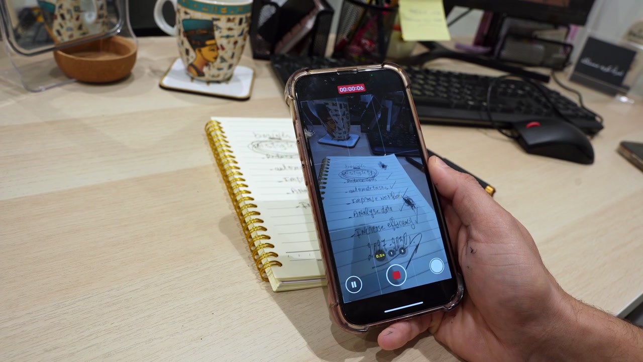 A close-up POV shot of a hand holding a smartphone to film a handwritten spiral notebook on a light wooden office desk. The recording screen shows business tasks alongside a coffee mug and keyboard.