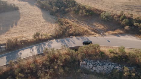 Aerial-drone-following-shot-over-a-black-sedan-passing-through-a-winding-road-in-El-Torcal-de-Antequera,-Sierra,Spain-on-a-sunny-evening