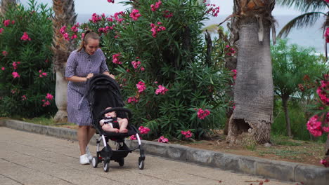 amidst a park in full bloom during the summer, a young mother enjoys a peaceful stroll with her baby in a stroller. her happiness is evident as she walks alongside her son