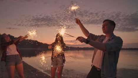 alegres amigos masculinos y femeninos están corriendo a lo largo de la playa al atardecer sosteniendo fuegos artificiales brillantes y luces fugitivas en cámara lenta. baile y fiesta de atardecer en la playa