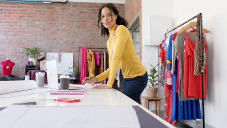 retrato de una feliz diseñadora de moda biracial de pie en la mesa trabajando en un estudio, en cámara lenta