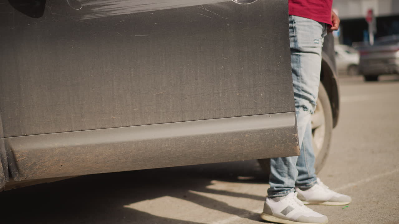 black man in red shirt exiting vehicle into bustling city lot, delivery driver vibe in jeans and white sneakers strides past dusty SUV, scans traffic and storefronts with focused posture, urban hustle