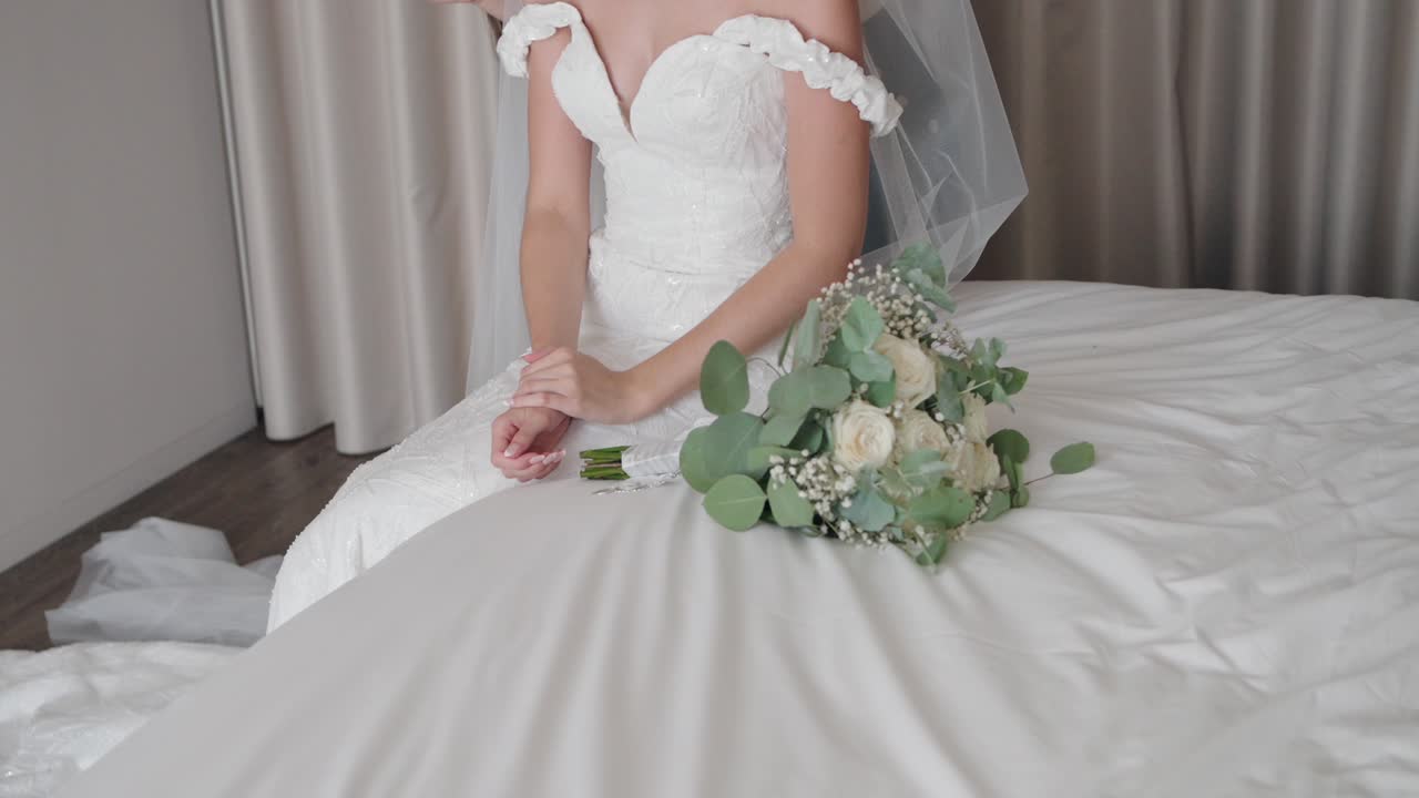bride sits on a bed, her white dress flowing, with a bouquet of white roses and greenery beside her