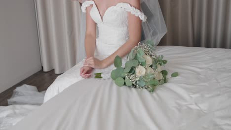 bride sits on a bed, her white dress flowing, with a bouquet of white roses and greenery beside her