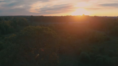 Slow-aerial-flight-over-trees-near-nepean-city,-ontario-at-sunset-with-the-sun-shining-over-the-hill-and-trees-and-a-highway-in-the-background