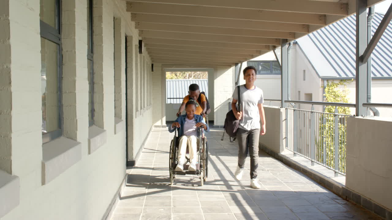 Multiracial students walking in school hallway, one pushing friend in wheelchair, copy space