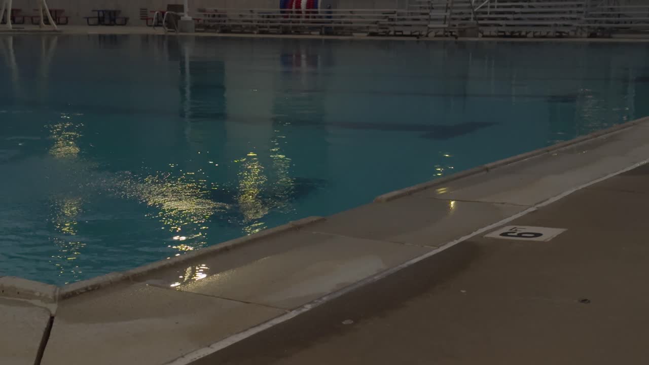 Wide shot of calm pool at night reflecting lights, with visible lane lines and 9 ft depth marking