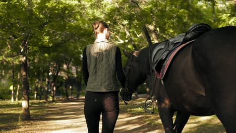 Back-view-of-young-brunette-jockey-girl-walking-with-horse-in-the-park-during-sunny-day