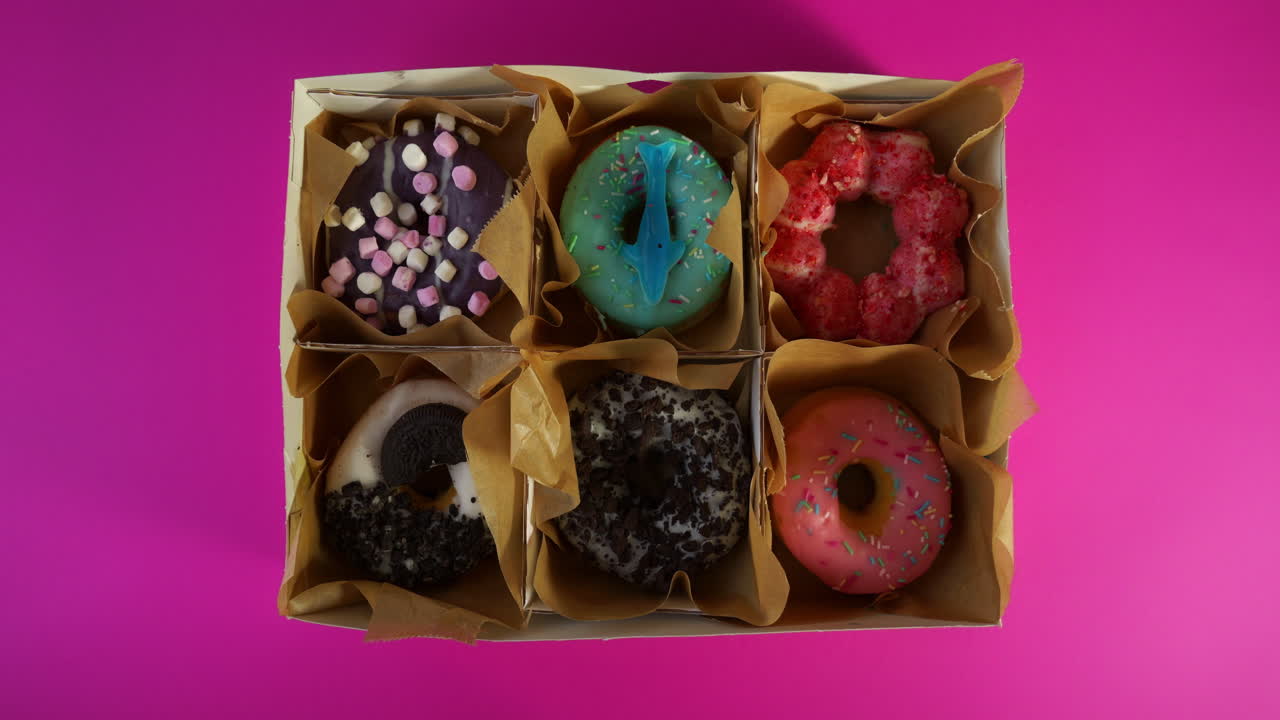 Overhead shot of hands gently placing a box of six assorted colorful donuts onto a bright pink background, showcasing vibrant toppings and a playful dessert arrangement.