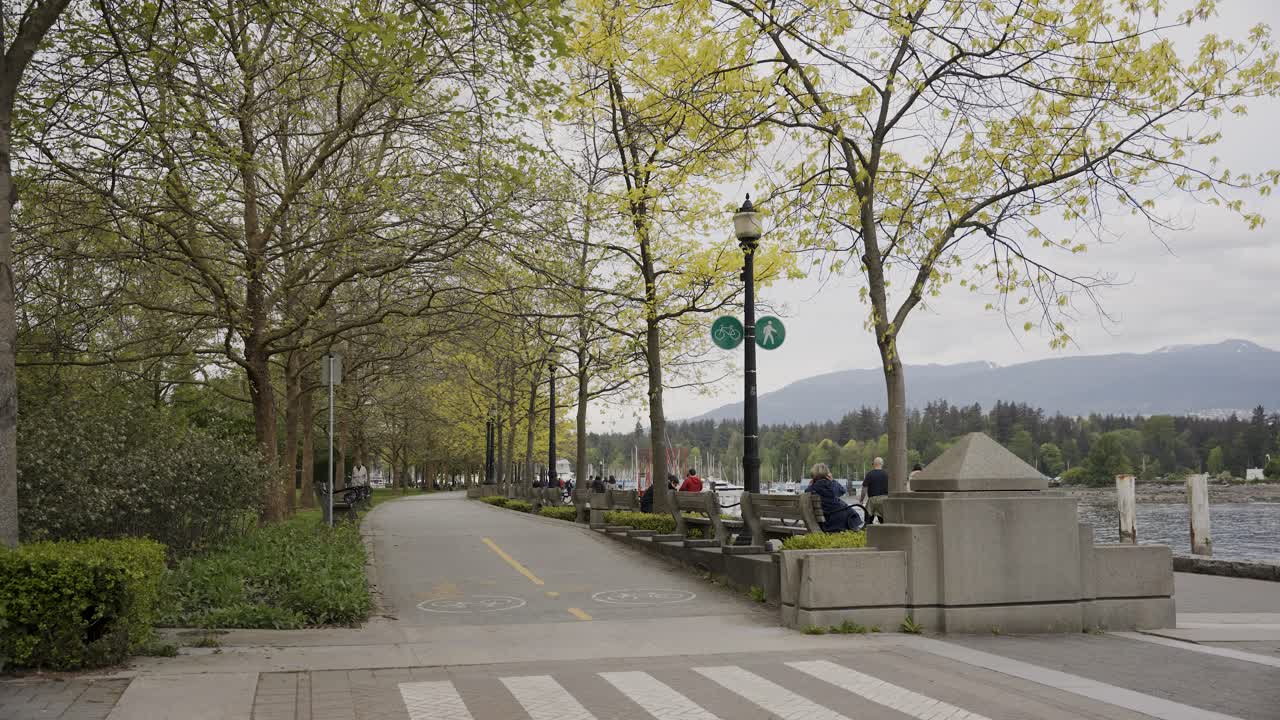 Orang-orang berjalan di promenade dekat jalur sepeda yang dikelilingi pepohonan, teluk Pusat Kota Vancouver, British Columbia, Kanada