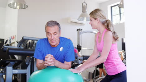 un anciano haciendo ejercicio con la pelota suiza alentado por un entrenador personal en el gimnasio.
