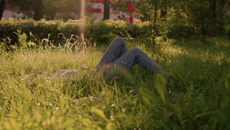 individual lying on grassy field under warm sunlight, legs bent, hands resting on body, surrounded by lush greenery, tall grass, and vibrant trees in background