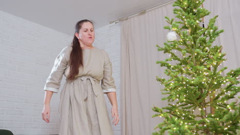woman in casual gown standing beside christmas tree while decorating with twinkling lights, carefully arranging glowing ornaments on lush green branches to create cozy warm festive atmosphere indoors