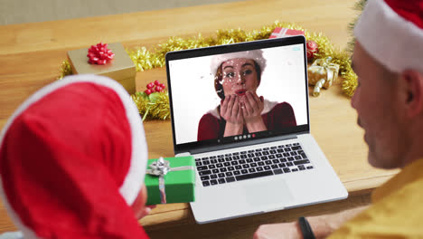 Padre-E-Hijo-Caucásicos-Con-Gorros-De-Papá-Noel-Usando-Una-Computadora-Portátil-Para-Una-Videollamada-Navideña-Con-Una-Mujer-En-La-Pantalla