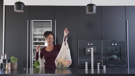 happy biracial woman putting down bags of grocery shopping and smiling in kitchen, in slow motion