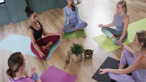 Multiracial-women-practicing-meditation-in-lotus-position-around-plant-and-singing-bowl