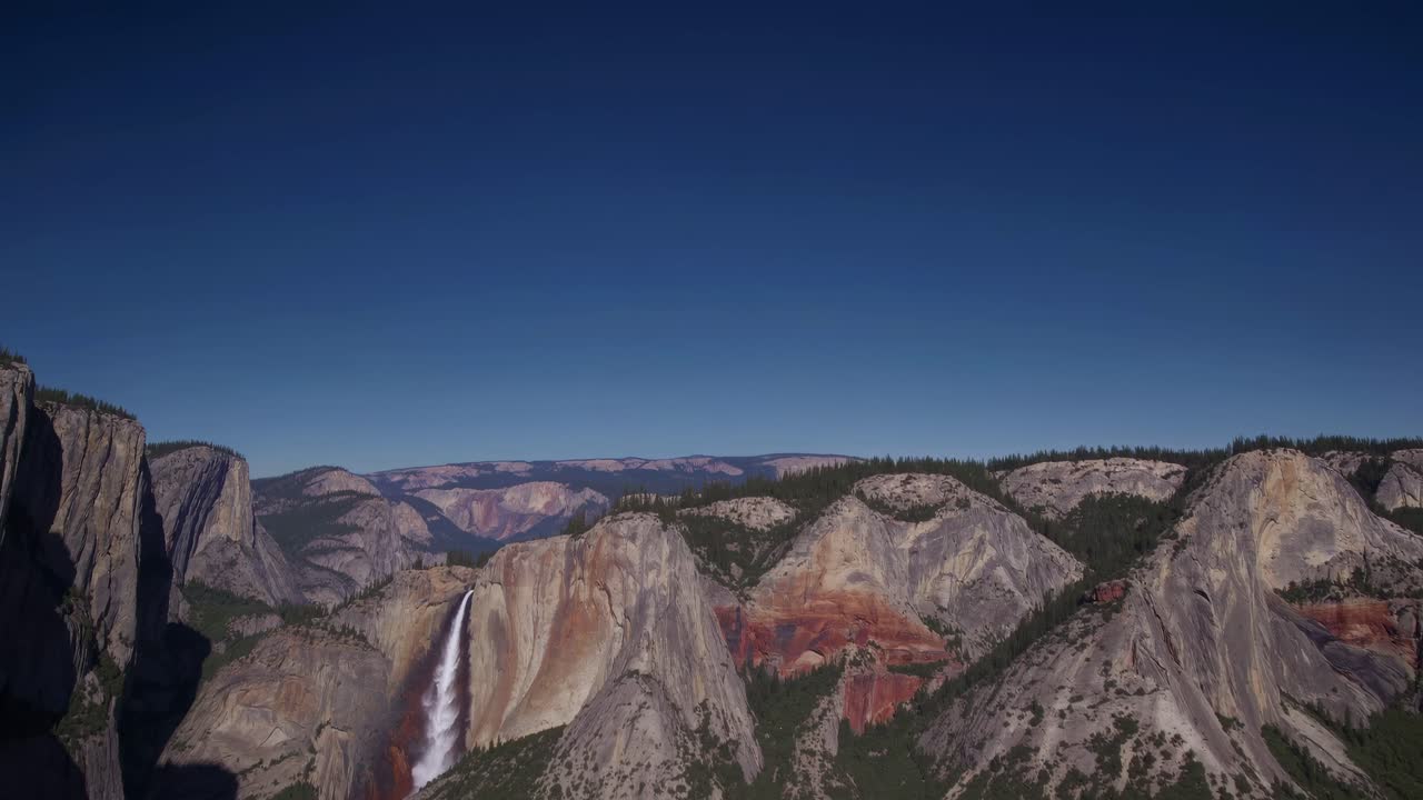 Aerial video capturing majestic mountains and a waterfall under a clear blue sky