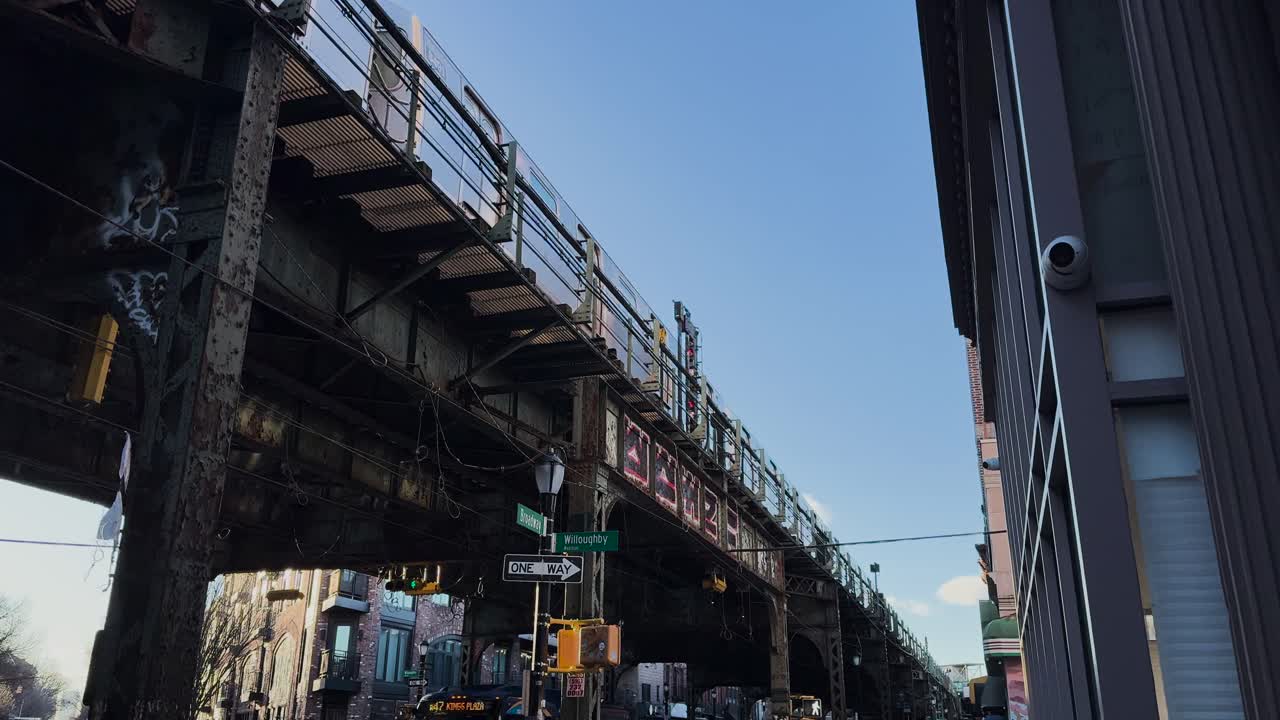 An above-ground subway train passes on an elevated track in Brooklyn. The camera pans down to show street life below, including traffic, pedestrians, and graffiti-covered urban textures.