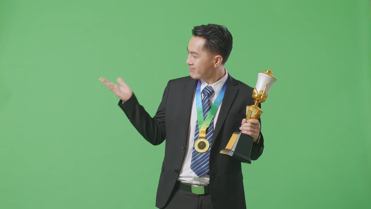 수트와 타이를 입은 아시아 사업가, 금메달을 들고 손에 금 트로피를 들고 미소 짓고 스튜디오의 녹색 화면 배경에 을 가리키고