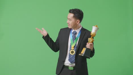hombre de negocios asiático en traje y corbata con una medalla de oro mirando un trofeo de oro en sus manos, sonriendo, y señalando hacia un lado en el fondo de la pantalla verde en el estudio