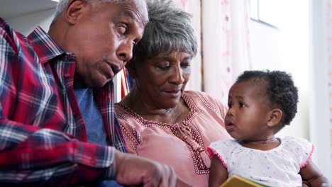 grandparents sitting on sofa reading book with baby granddaughter at home