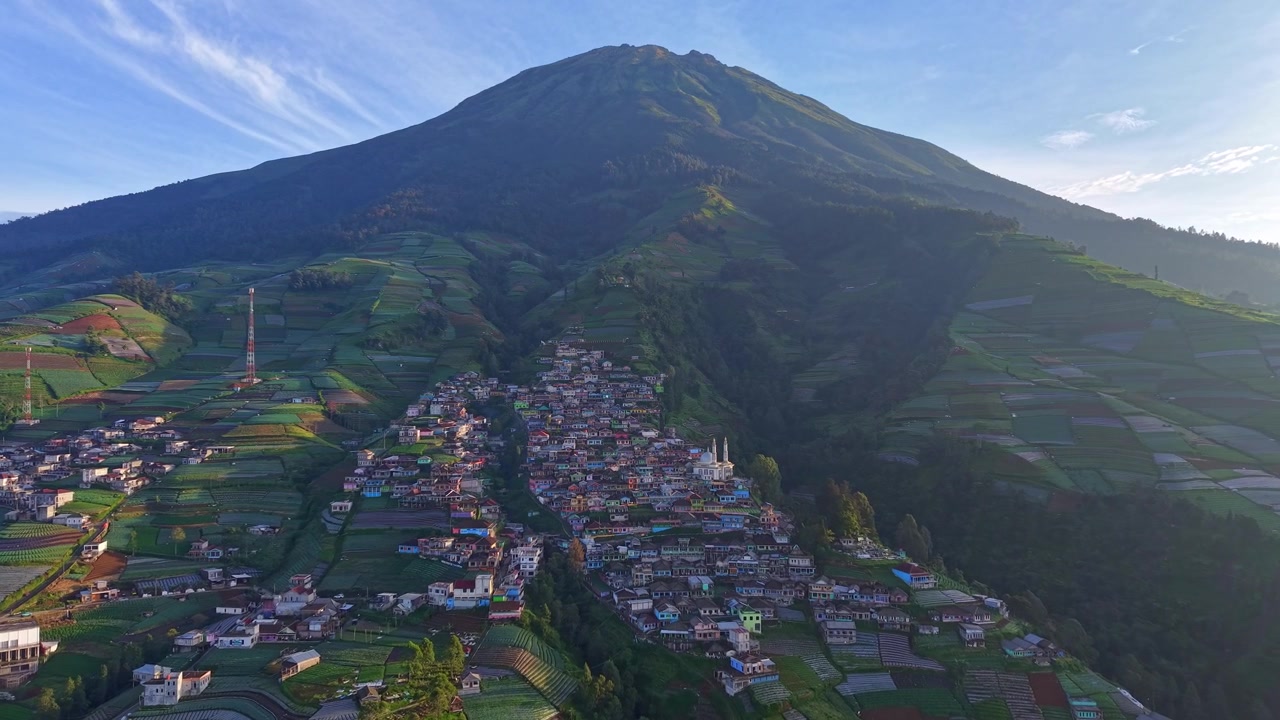 Vista aerea di un villaggio di montagna circondato da verdi colline e terreni agricoli sotto un cielo azzurro e limpido. Paesaggio rurale suggestivo con uno sfondo montuoso spettacolare e un'atmosfera di campagna tranquilla.
