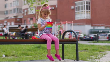 Fröhliches-Mädchen,-Das-Mit-Pop-It-Spielt,-Einem-Einfachen-Sensorischen-Anti-Stress-Spielzeug-Mit-Grübchen-Auf-Einer-Spielplatzbank