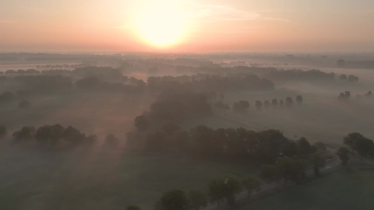 Aerial video captures serene sunset in Twente, Netherlands, with misty clouds and gentle light. Features expansive fields and trees, creating a tranquil atmosphere with golden hues