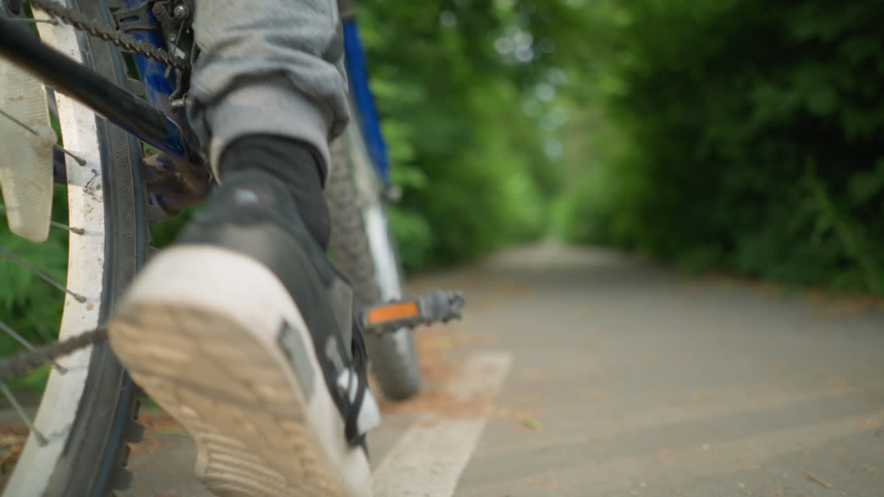 자전거의 페달을 발로 뒤로 는 누군가의 클로즈업, 설정은 도로 표시와 함께 울창한 초록색의 흐릿한 배경으로 포장 된 경로입니다.
