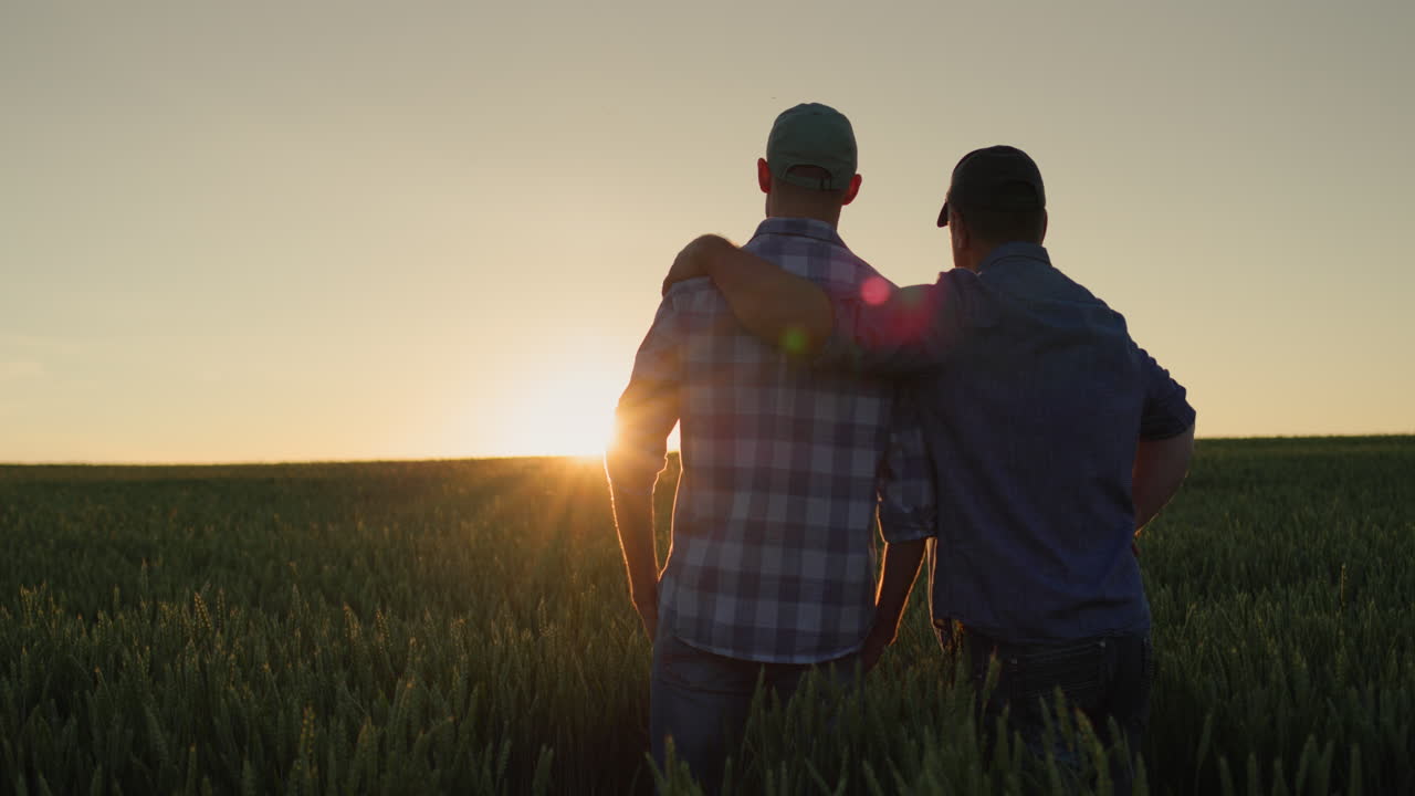 Padre contadino che abbraccia il figlio adulto e guarda il tramonto sul campo insieme.