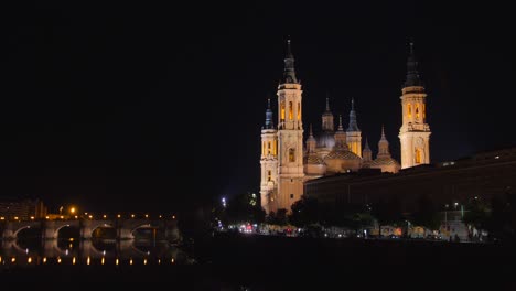 Tráfico-Conduciendo-A-Lo-Largo-Del-Río-Ebre-Pasando-Por-La-Catedral-basílica-De-Nuestra-Señora-Del-Pilar-Con-El-Puente-De-Piedra-Por-La-Noche-En-Zaragoza,-España