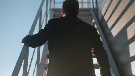 back view individual climbing staircase changing briefcase left hand to right hand with light reflecting on iron railing, slow motion upward motion