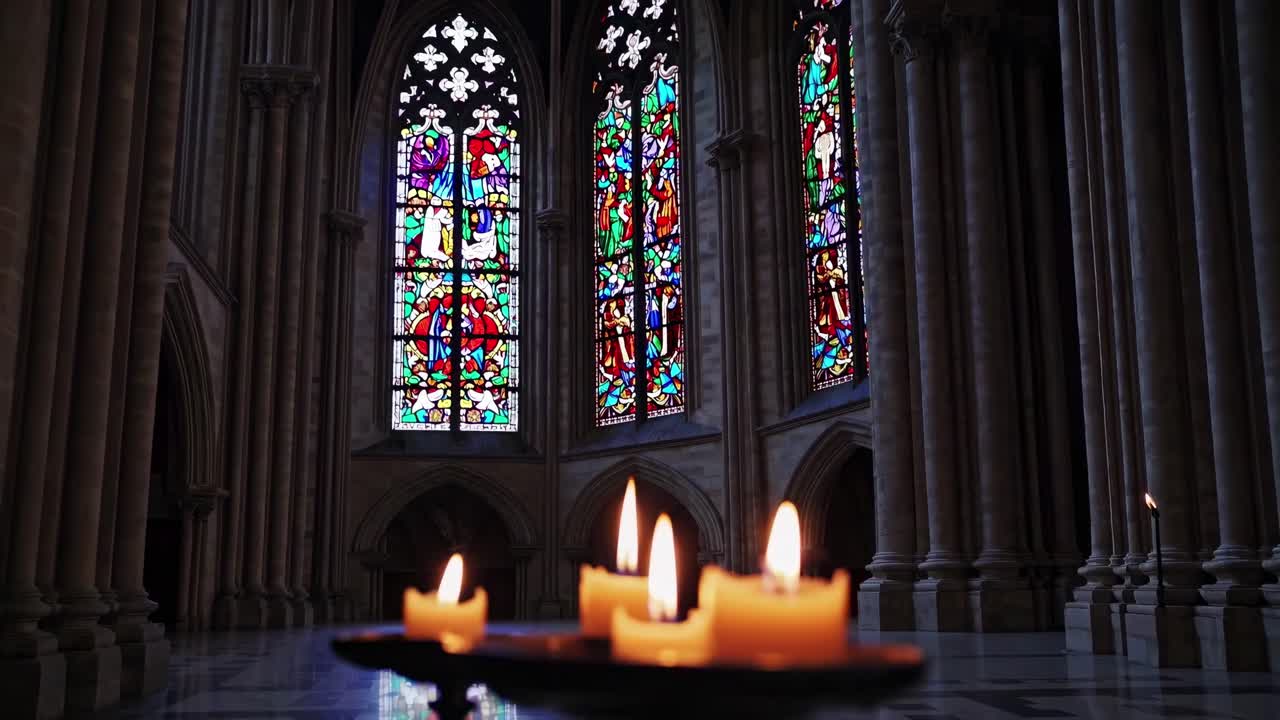 Low-angle video of lit candles in a dim cathedral, highlighting colorful stained glass windows