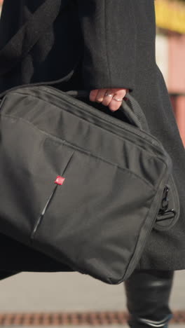vista lateral de una mujer que lleva una bolsa negra con la mano adornada con anillos, caminando con confianza, vestida con ropa negra con edificio rojo borroso y vegetación decorativa en el fondo