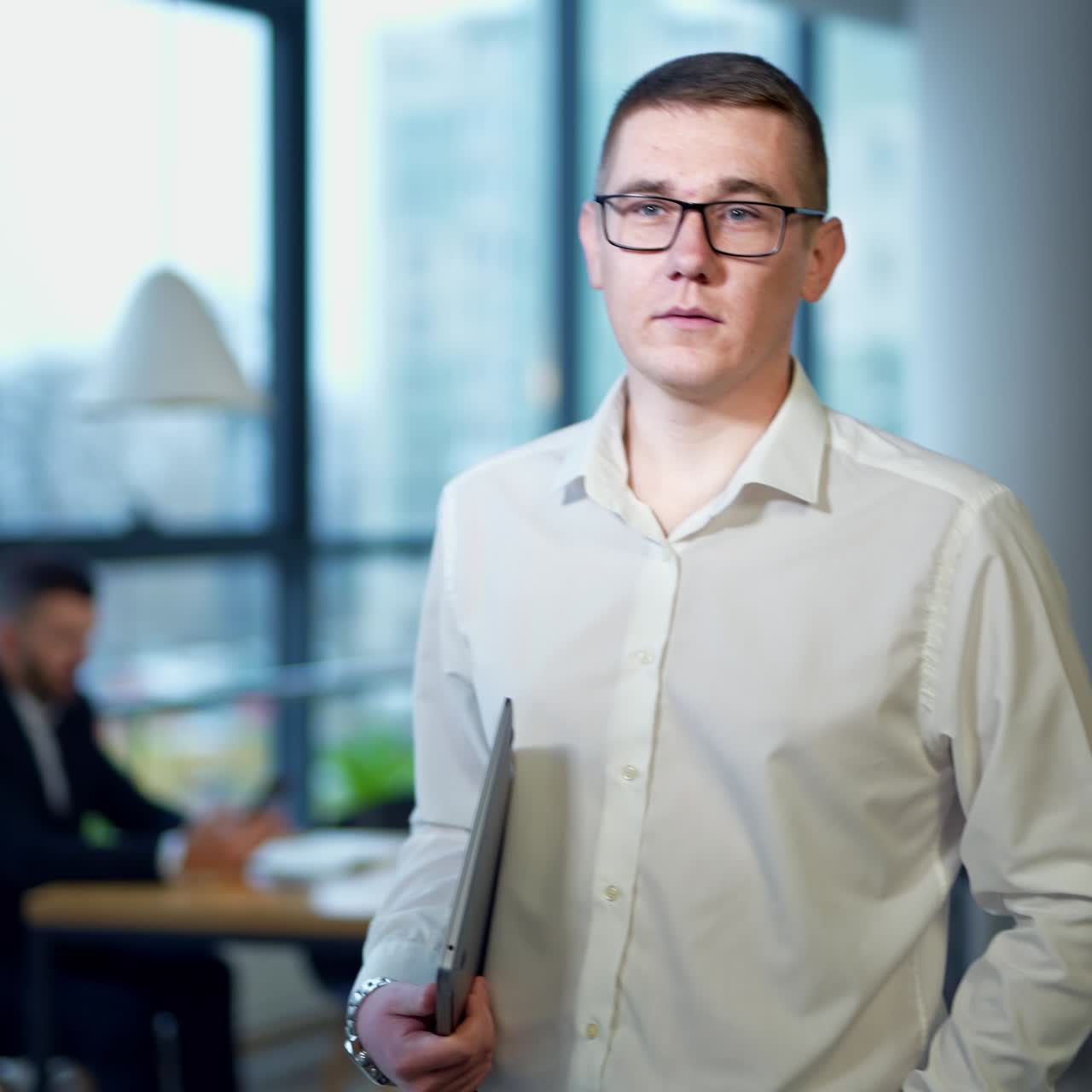 Caucasian man wearing glasses and holding a laptop in the office. Male employee portrait standing against the panoramic windows backdrop