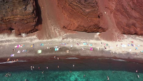 aerial top down flying sideways across red beach with turquoise water, tourists, mountains and red colored sand in santorini, greece