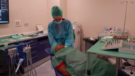 asistente de dentista cubriendo la cara del paciente antes de la cirugía mientras el médico se pone un traje de ppe