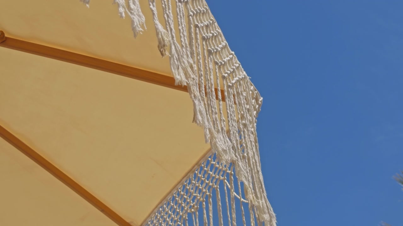 Bela tomada em 4K de ângulo baixo de guarda-sóis de macramê brancos com franjas em um resort de praia de luxo. Apresenta uma fileira organizada de espreguiçadeiras, palmeiras tropicais e um céu azul claro.