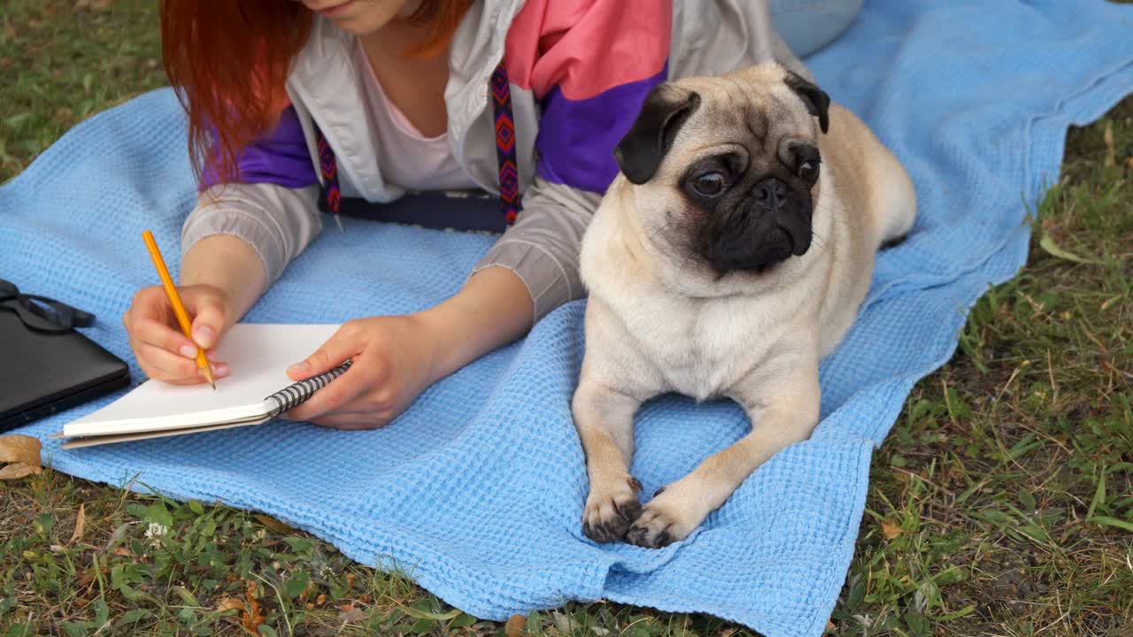 chica acostada en un césped y escribiendo, su pug acostado al lado