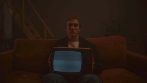 portrait of young man sitting on sofa, holding vintage tv with static screen