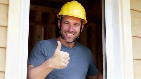 smiling builder with yellow helmet giving a thumbs up, stand near window.
