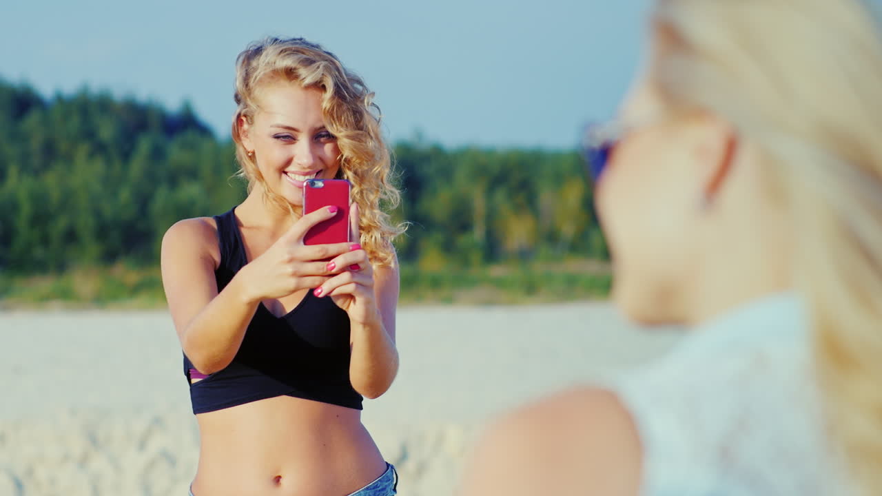 승인 제스처를 보여주는 해변에서 그의 여자 친구에게 스마트 폰으로 사진을 찍는 젊은 여자