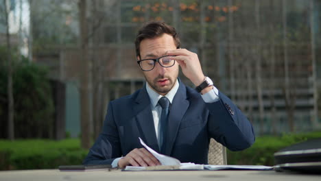 caucasian male executive working alone reviewing paperwork in outdoor office park, then taking off glasses and massaging forehead having eyes pain or headache