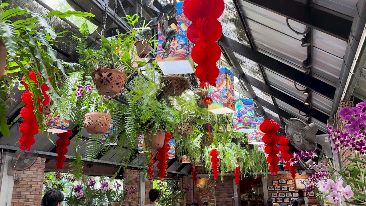 Colorful hanging plants, orchids, and red lanterns decorate a bright indoor garden market walkway