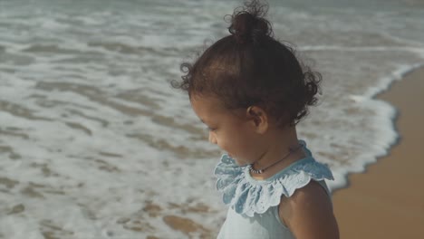 Semi-close-up-shot-of-Indian-child-in-light-blue-dress-watching-calm-waves-at-Benaulim-Beach,-Goa