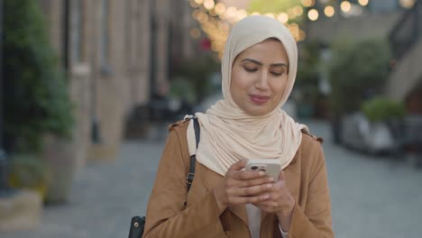 muslim woman wearing hijab walking along city street sending text message to date on mobile phone 1