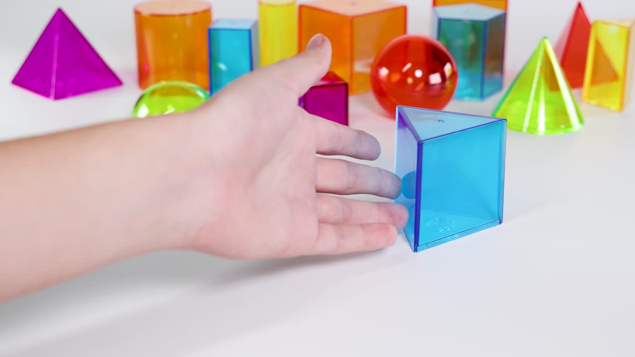 Hands manipulate vibrant 3D geometric shapes on a white surface, highlighting educational engagement with prisms, spheres, and pyramids